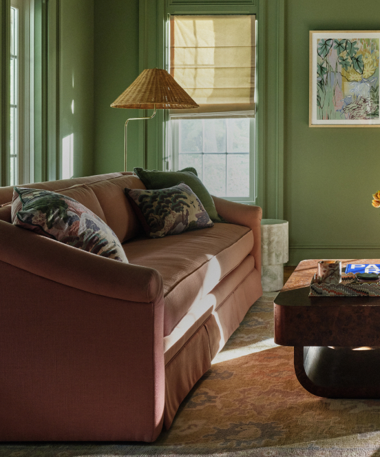 Green-walled living room with brown velvet sofa, decorative pillows, wicker floor lamp, and framed botanical artwork.