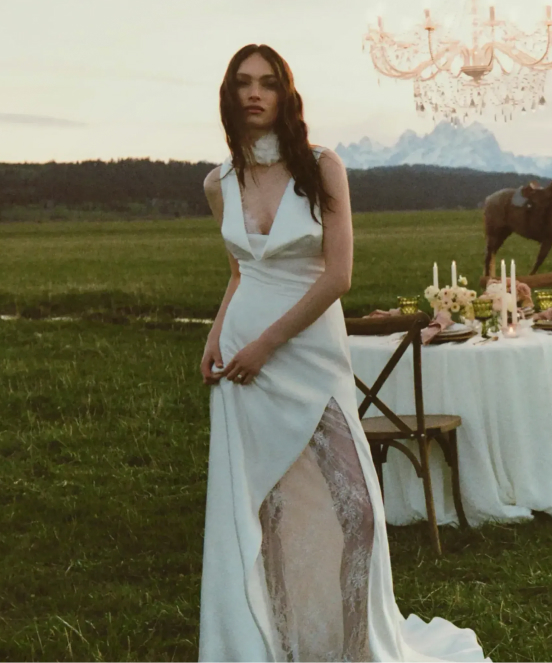 Woman in white wedding dress with lace detail standing in field with outdoor dining table, chandelier, and horse in background
