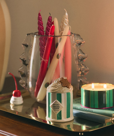 Decorative candles in pink, red, and white arranged on a mirrored tray with a lit green striped candle and small ornament.