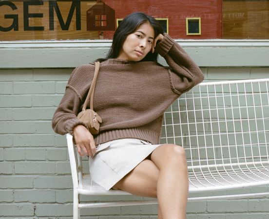 Woman in brown sweater and white skirt sitting on white metal bench against brick wall with "GEM" text visible