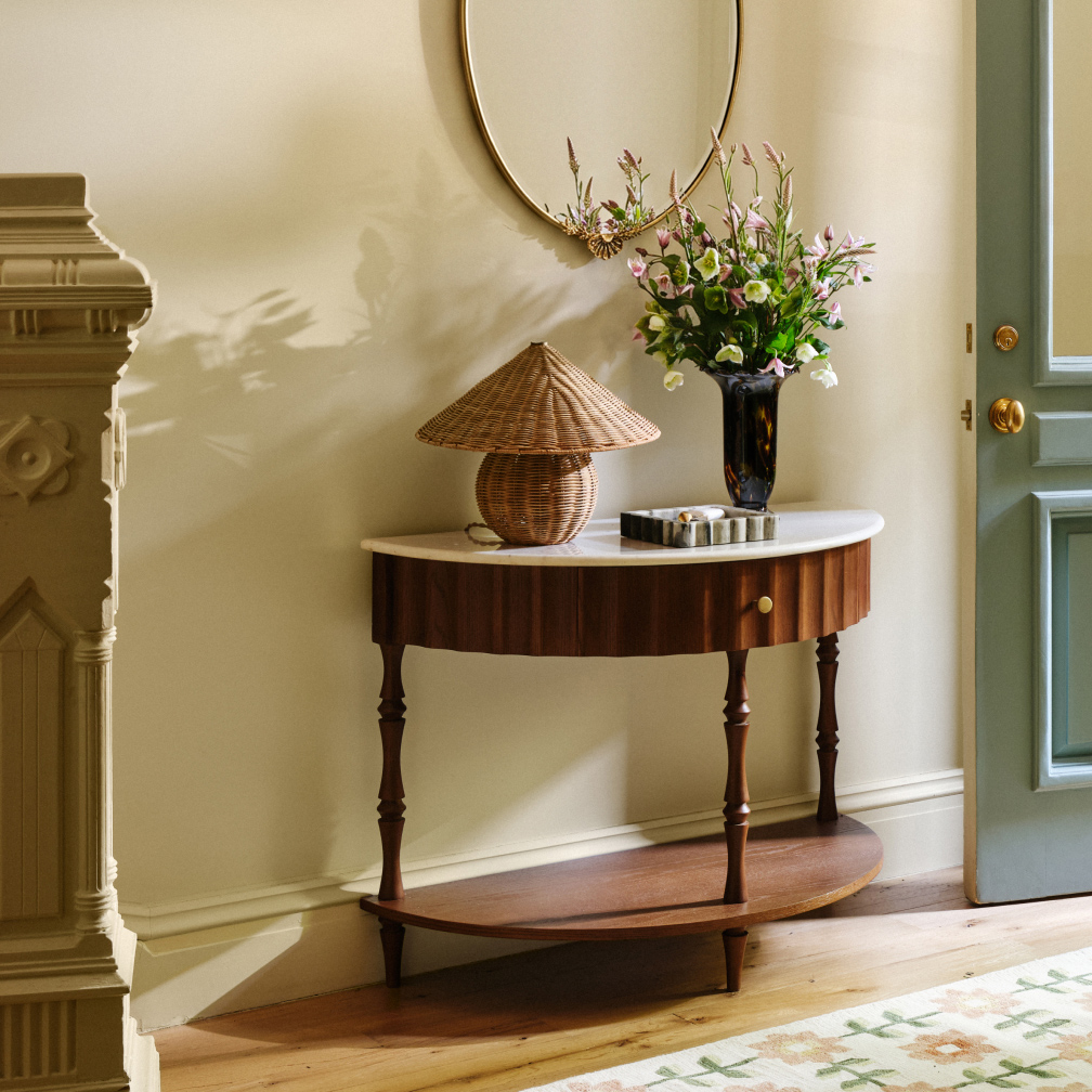 Elegant entryway with oval console table, wicker mushroom lamp, floral arrangement in dark vase, and round gold mirror.