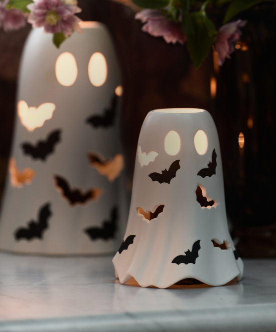 White ceramic ghost-shaped candle holders with glowing faces and black bat cutouts, with pink flowers in background.
