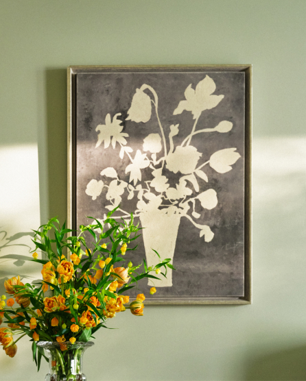 Framed floral artwork in gray tones on sage green wall with vase of orange and yellow flowers in foreground.