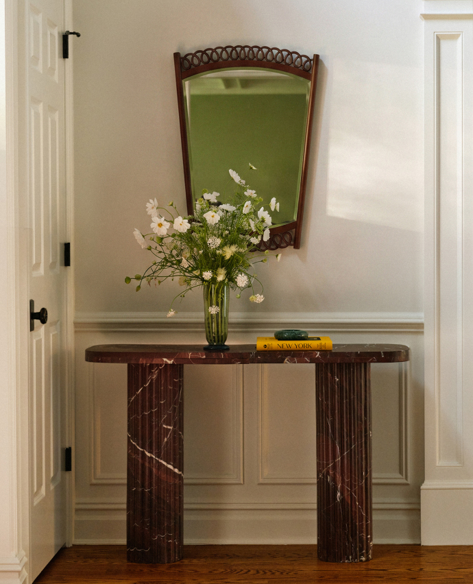 Elegant entryway with dark marble console table, white flower arrangement in green vase, decorative mirror, and books.