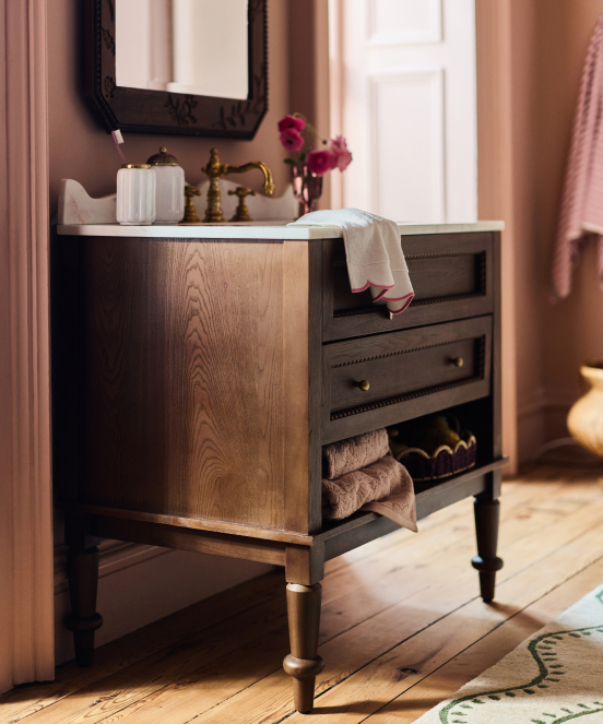 Wooden bathroom vanity with open drawer displaying towels, brass faucet, white canister, pink flowers, and mirror above.