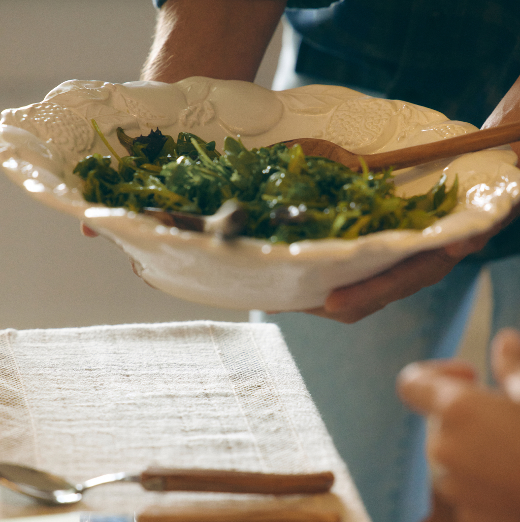 Person holding white ceramic bowl with leafy green salad and wooden serving spoon over a table with white linen.