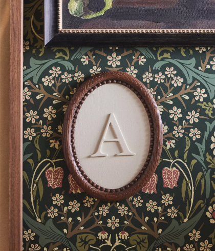 Ornate oval frame with embossed letter A on cream background, displayed on dark green wallpaper with yellow floral pattern.