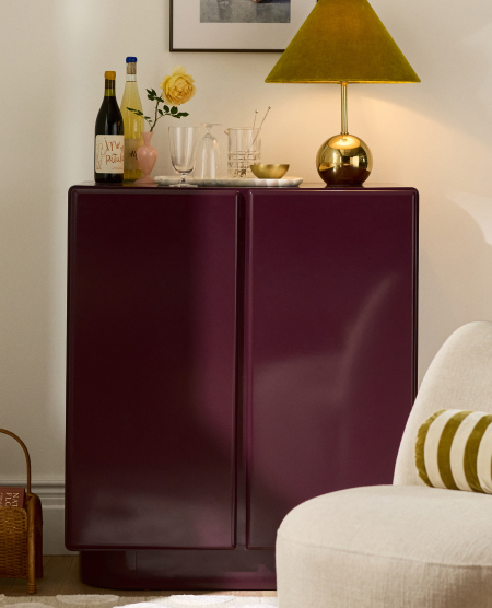 Burgundy cabinet with brass lamp featuring olive green shade, wine bottle, glassware, and yellow flowers on top
