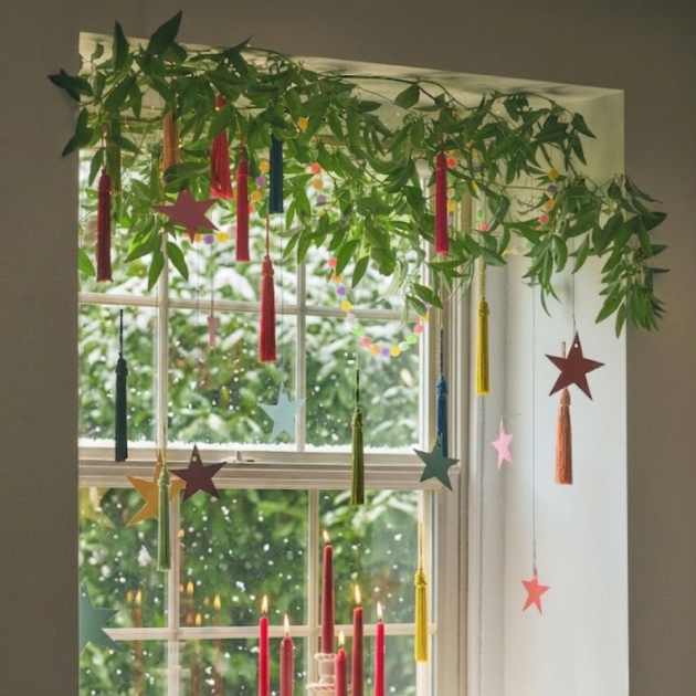 Window decorated with green garland, colorful hanging candles, and star ornaments with outdoor lights visible through glass