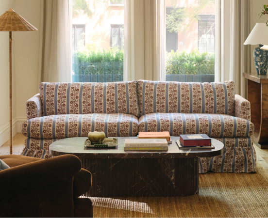 Living room with patterned sofa, dark oval coffee table with books, wicker lamp, and large windows overlooking greenery.