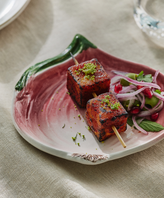 Two grilled meat skewers with herbs on a pink plate, accompanied by a fresh salad with red onions and a green pepper.