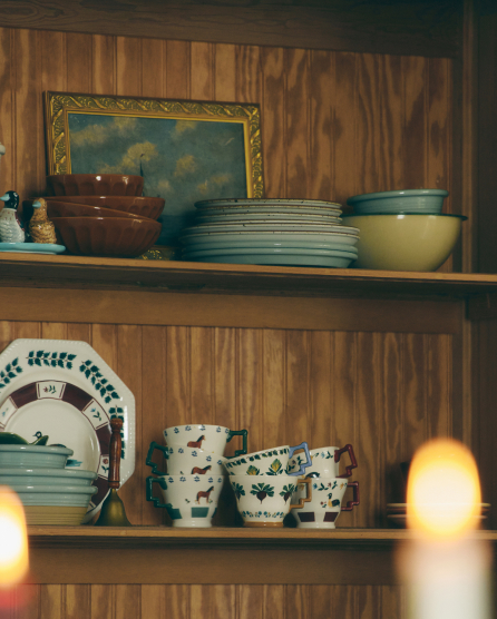Wooden shelves displaying vintage dishware including stacked plates, bowls, and decorative cups with floral patterns and a framed painting.