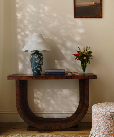 Curved wooden console table with blue ceramic lamp, flower vase, and books against cream wall with dappled sunlight shadows.