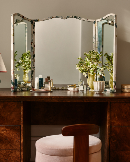Vintage tri-fold vanity mirror on wooden desk with pink velvet stool, decorated with green plants and beauty products.