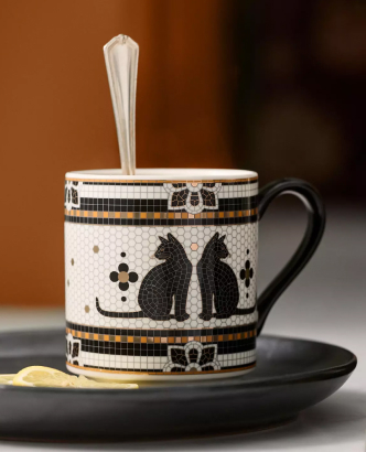 White ceramic mug with black cat silhouettes and decorative border pattern, sitting on black saucer with spoon inside.