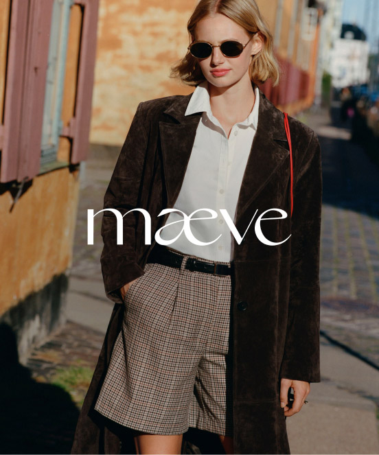 Woman in sunglasses wearing dark coat, white shirt, and plaid skirt walking on urban street with colorful buildings.
