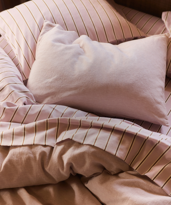 Soft pink pillow on pink and gold striped bedding with rumpled beige blanket in warm natural lighting