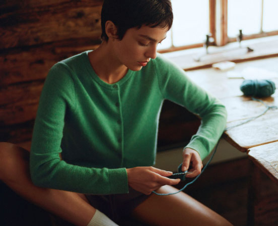 Woman in green sweater looking down at smartphone while sitting indoors near a window with natural light