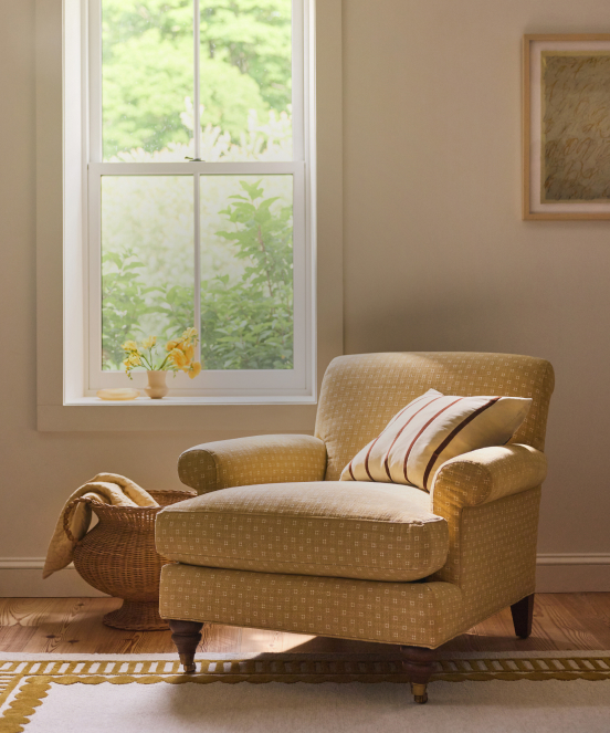 Cozy beige armchair with striped pillow beside a white-framed window overlooking green foliage in a neutral-toned room.