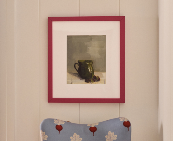 Framed still life painting of a green pitcher in red frame above blue floral pillow with text Art & Mirrors below
