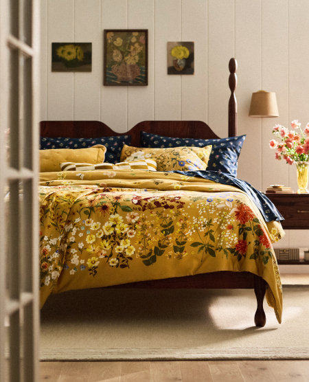 Bedroom with dark wood bed featuring golden yellow floral bedding, navy tufted headboard, white paneled walls with vintage art, and bedside lamp.