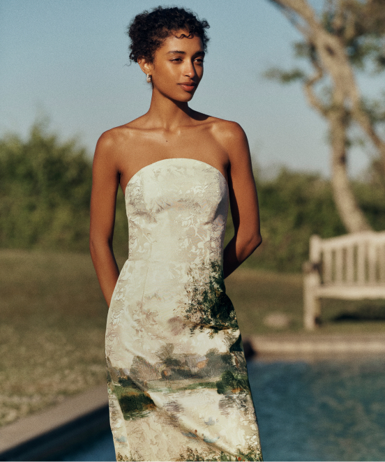 Woman in strapless white dress with nature print standing outdoors by pool with palm trees in background