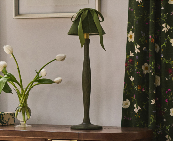 Green leaf-shaped table lamp on wooden surface next to white tulips in vase with floral curtain in background
