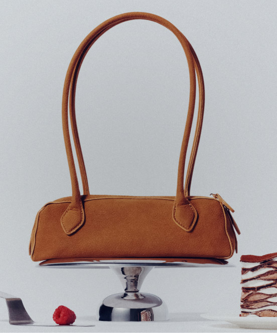 Brown leather handbag with long handles displayed on a silver pedestal against a light gray background.