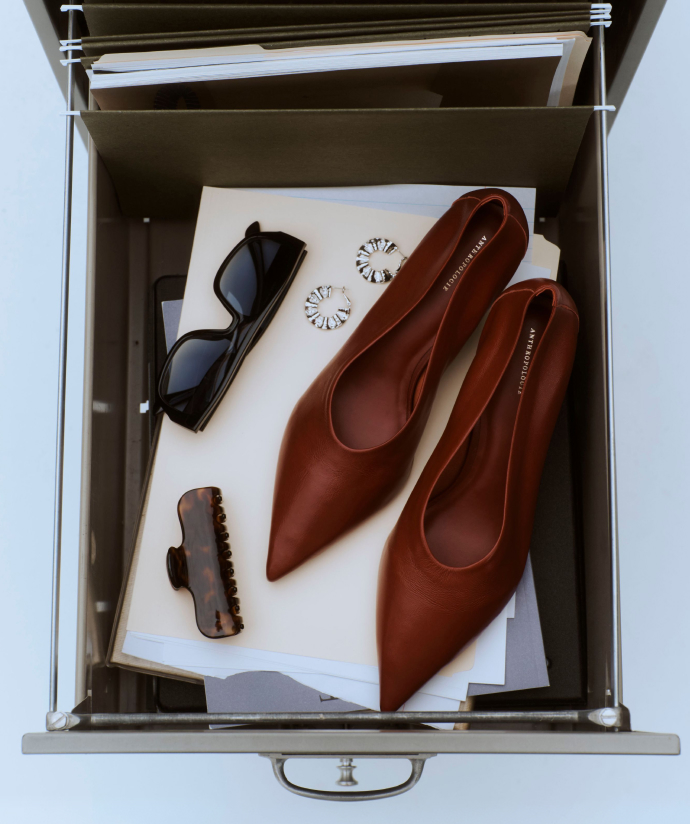 Open drawer containing brown leather pointed-toe flats, black sunglasses, silver flower earrings, and a brown hair clip.