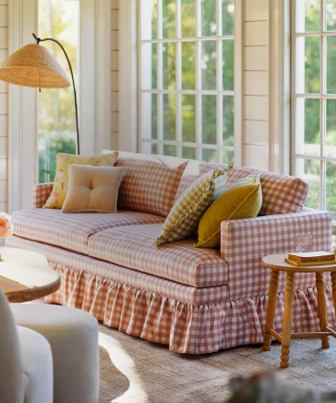 Pink gingham sofa with ruffled skirt and colorful pillows in a bright sunlit room with large windows and a woven floor lamp