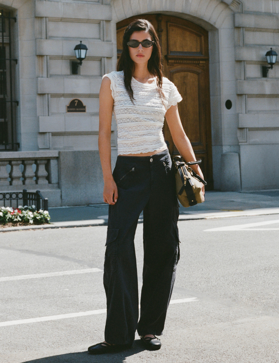 Woman in sunglasses wearing white textured crop top and black cargo pants stands on street in front of stone building.