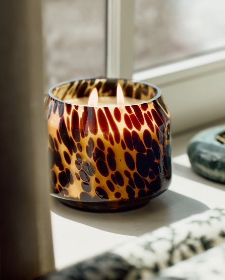 Lit candle in a glass holder with tortoiseshell pattern in amber, brown, and black tones on a white windowsill.