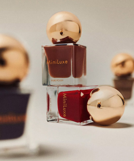 MiniLuxe nail polish bottles with gold spherical caps, one brown and one red, arranged on a neutral background.