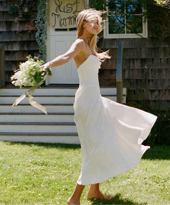 Bride in white dress twirling on lawn with bouquet, hair flowing, rustic building with "Just Married" sign in background