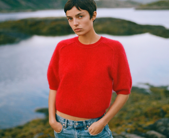 Woman in red sweater and jeans standing by rocky coastline with water in background