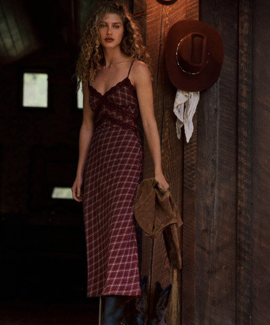 Woman in burgundy plaid slip dress stands by rustic wooden wall with brown hat and white cloth hanging beside her.