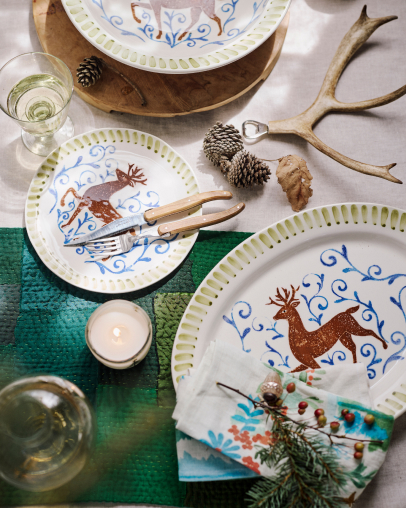 Festive table setting with decorative plates featuring reindeer designs, natural antlers, pinecones, candles, and evergreen accents.