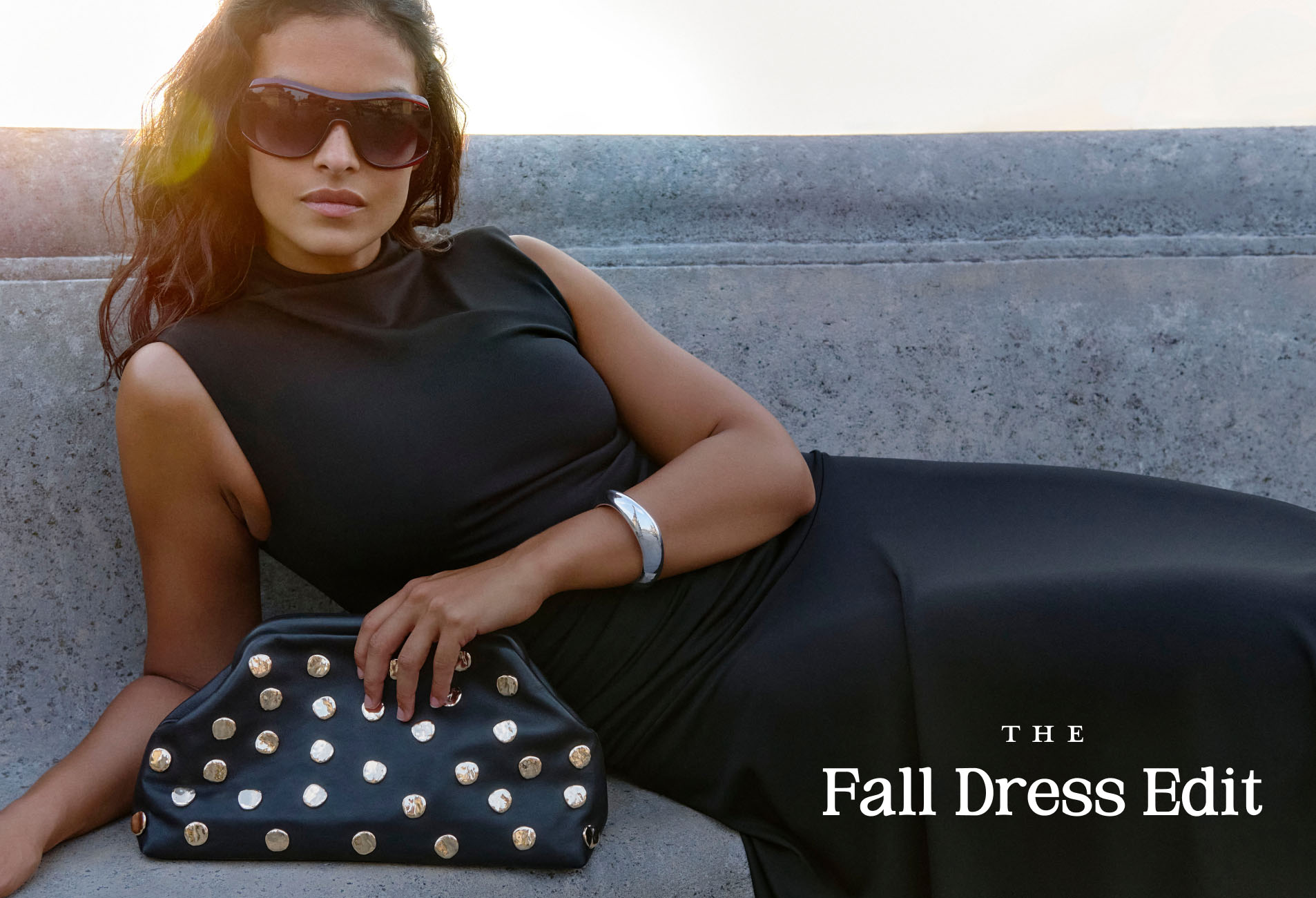 Woman in black outfit and sunglasses sitting on concrete steps with studded handbag. Text reads "THE Fall Dress Edit"