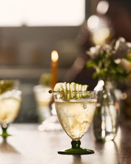 Cocktail in green-stemmed glass with white chocolate drizzle rim, lit candle and flowers in soft-focus background