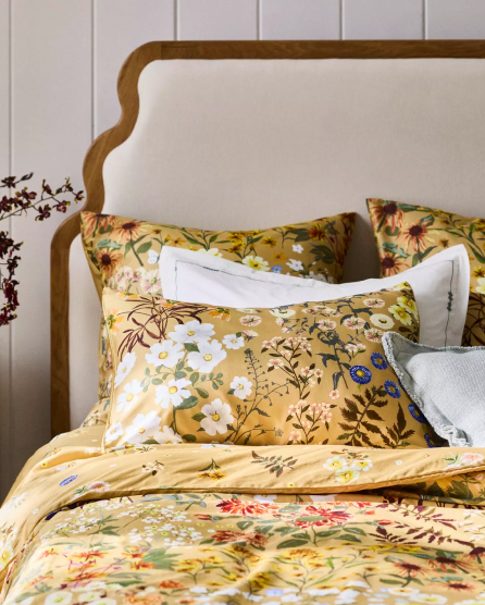 Bed with golden yellow floral bedding featuring botanical prints, white accent pillows, and an upholstered headboard with wood trim.