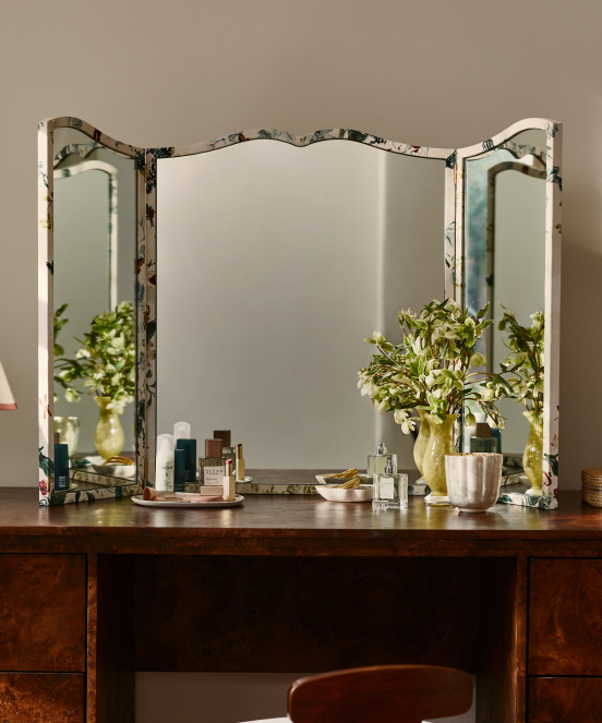 Vintage tri-fold vanity mirror with distressed white frame on wooden dresser, flanked by green plants in vases and beauty products.