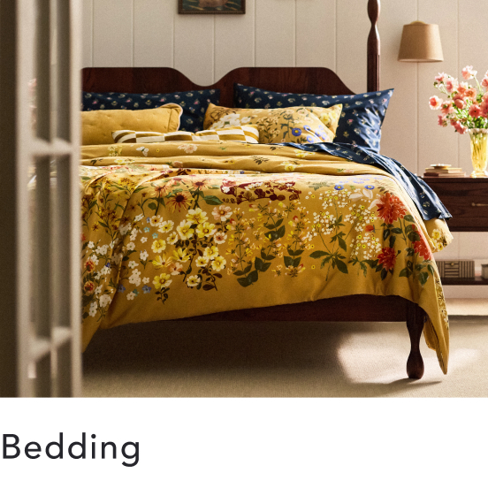 Wooden bed with golden yellow floral bedding and navy blue pillows in a bright bedroom with white walls and a bedside lamp