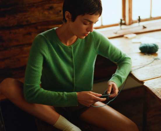 Woman in green sweater sitting on wooden floor looking down at smartphone in sunlit room with exposed brick wall
