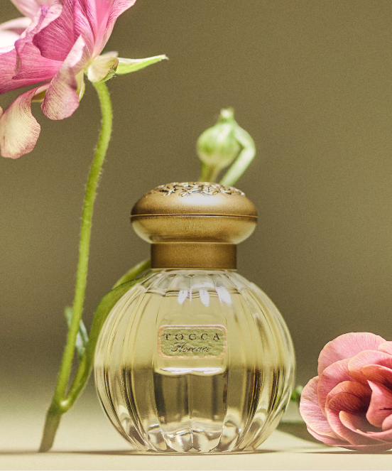 Tocca Florence perfume bottle with gold cap and ribbed glass design, surrounded by pink flowers on neutral background.