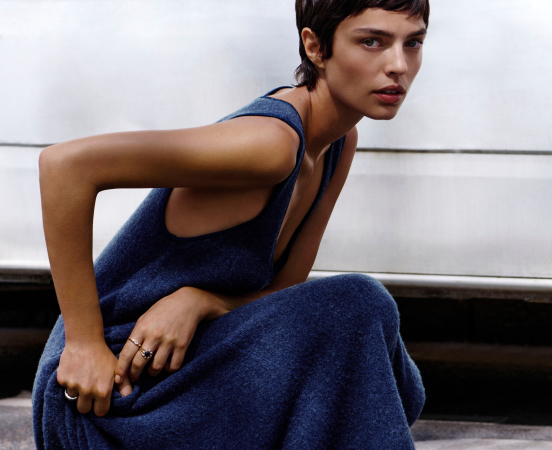 Woman with short dark hair wearing a textured navy blue sleeveless dress, seated against a white paneled wall.