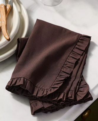 Dark brown cloth napkin with ruffled edges folded on white marble surface beside plates and wooden utensils