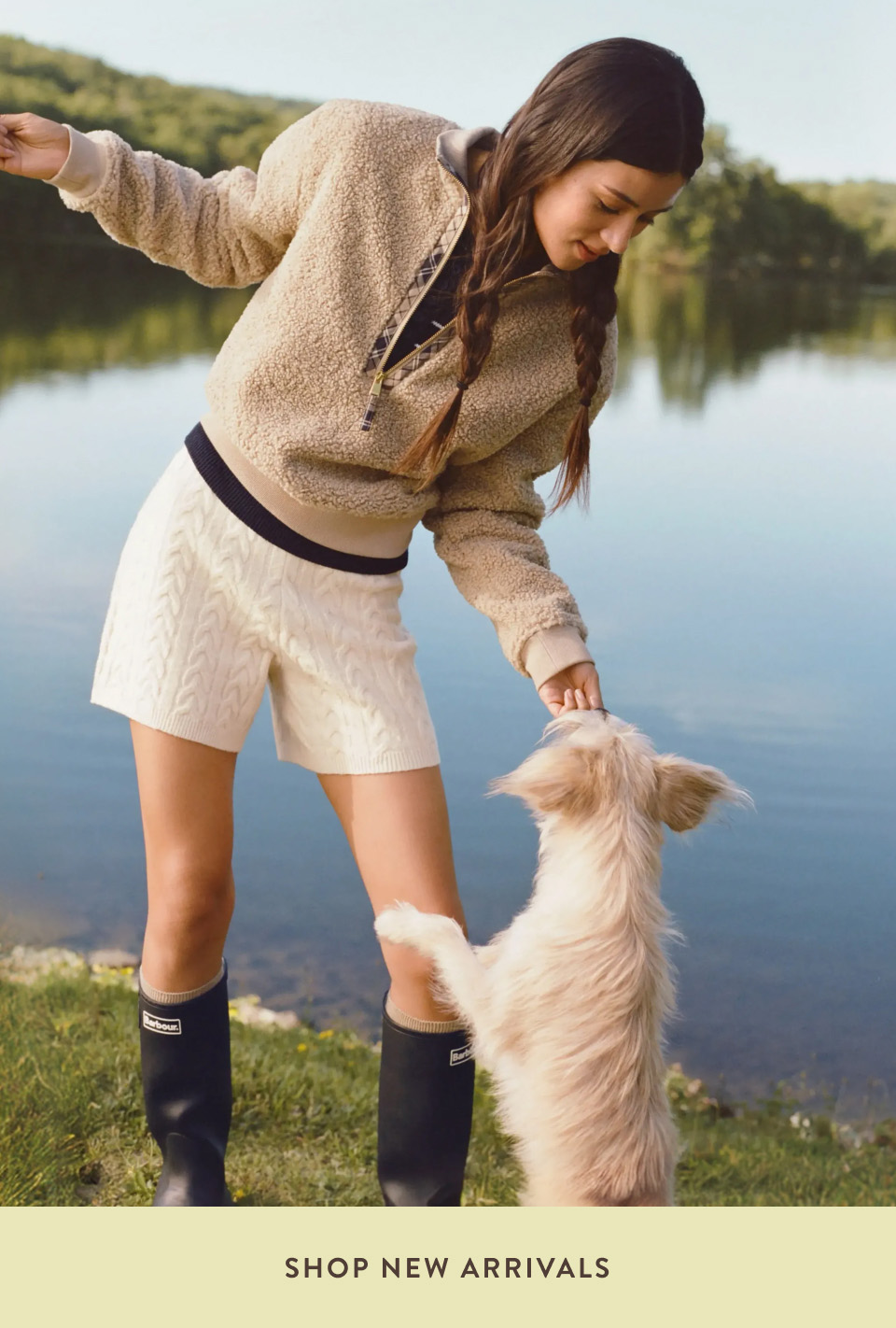 Woman in beige fleece jacket and cream shorts playing with fluffy dog by lake. Text reads SHOP NEW ARRIVALS.