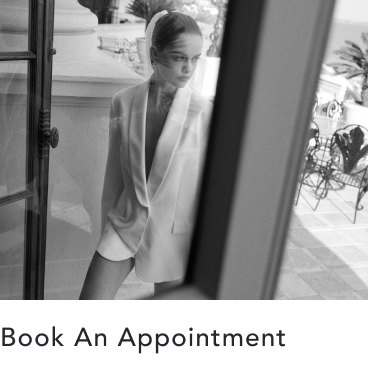 Black and white photo of woman in blazer and sunglasses standing in doorway. Text reads "Book An Appointment"