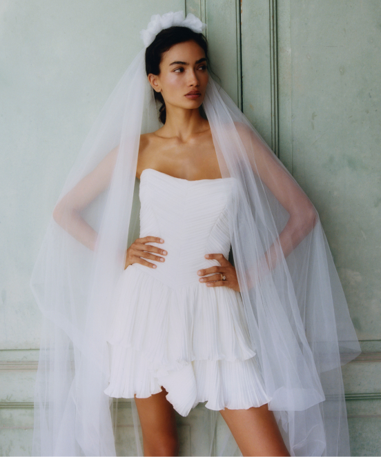 Bride in short white strapless tulle dress with flowing veil, posing against pale green paneled wall.