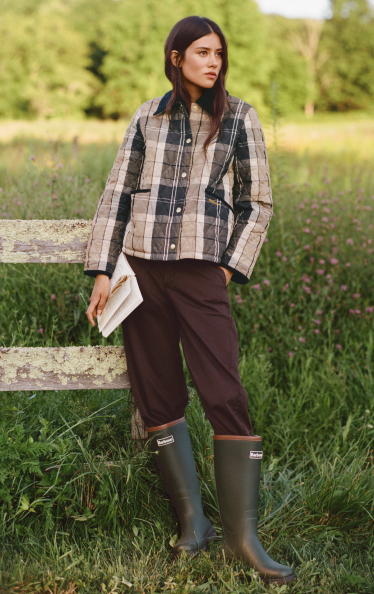 Woman in plaid quilted jacket, brown pants, and green rain boots standing by wooden fence in grassy field with wildflowers.